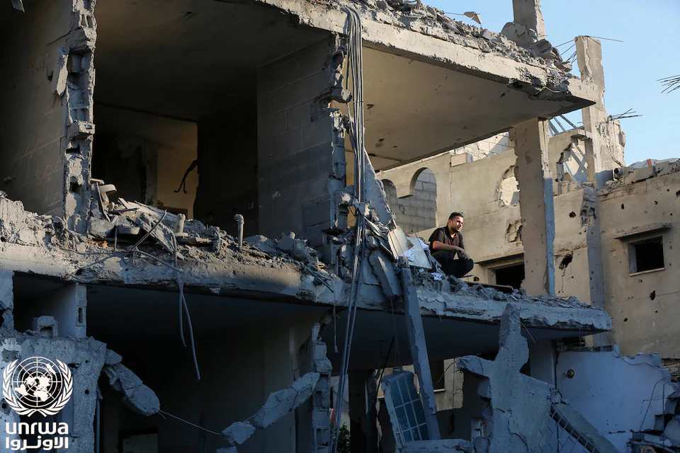 A man sits on the exposed floor of a heavily damaged multi-storey home surrounded by broken concrete and walls.