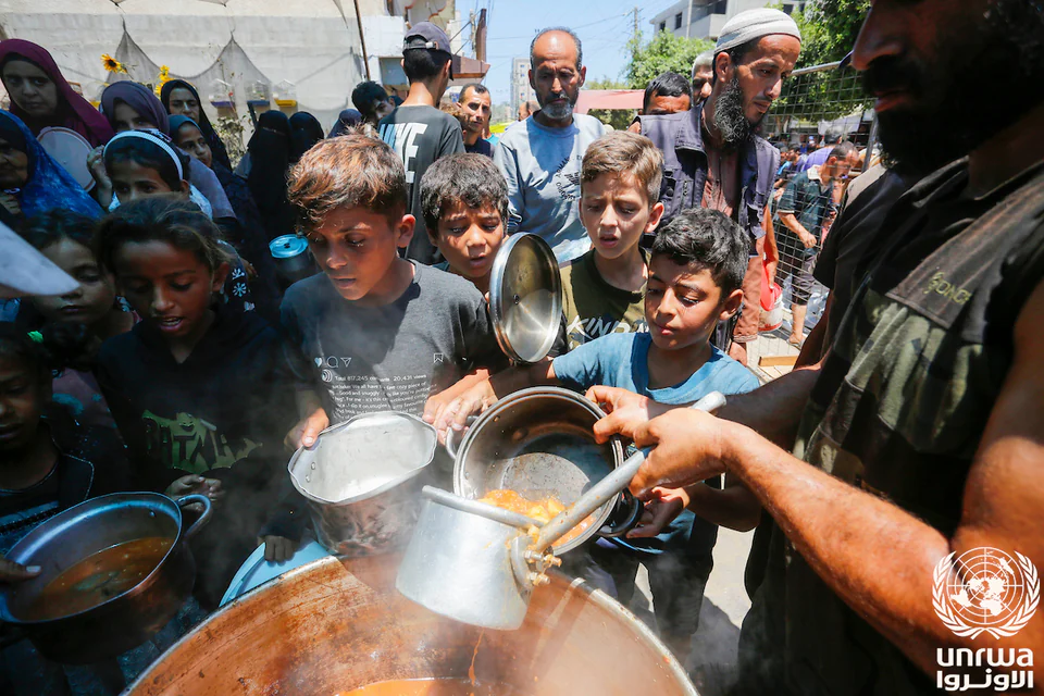 Adults and children gather around large cooking pots while food is served into metal containers outdoors.
