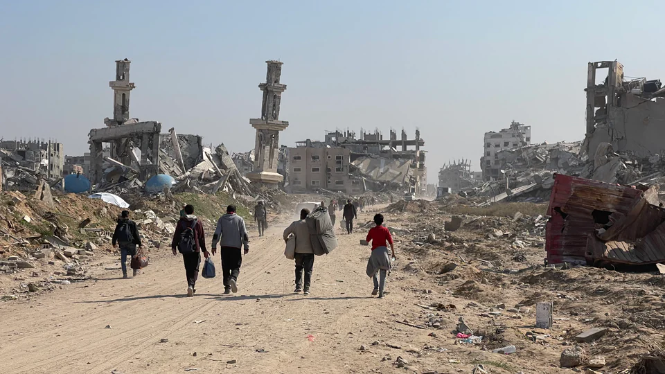Several people carrying bags and bedding walk along a dirt road between heavily damaged buildings in the Gaza Strip.